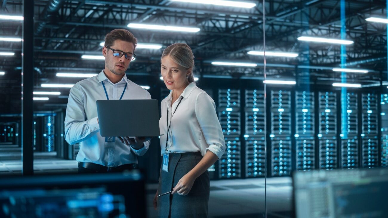2 Personen stehen vor einem Server und besprechen Informationen auf einem Laptop