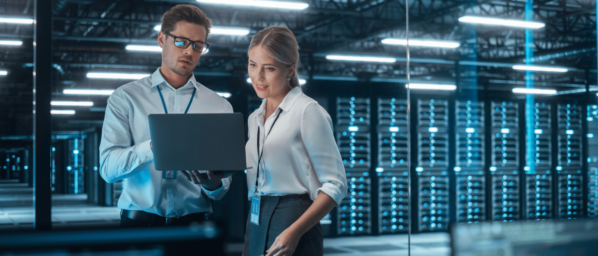 Zwei Technikexperte unterhalten sich über etwas auf einem Laptop. Der Hintergrund zeigt einen Serverraum.